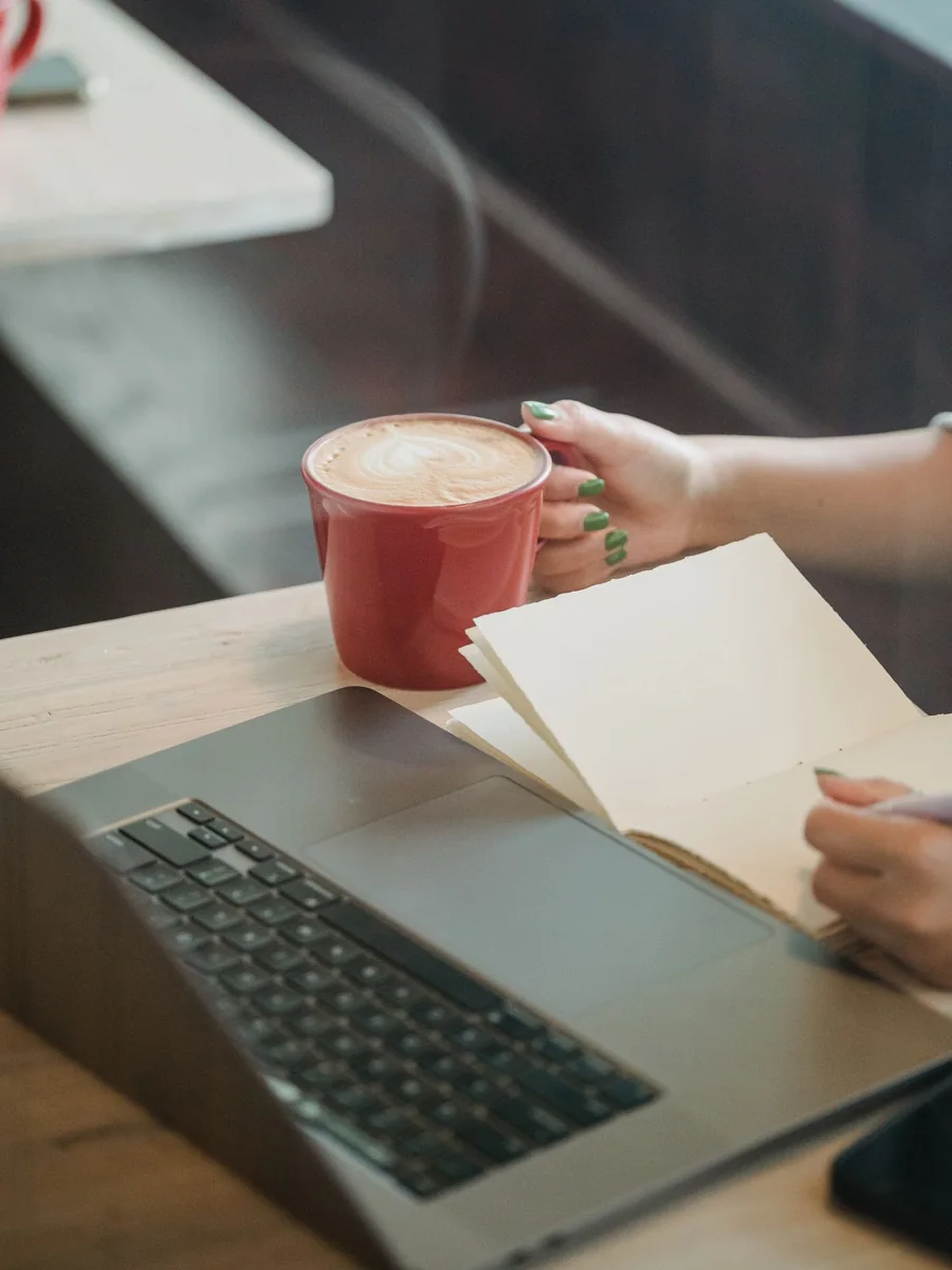 A person writing in a notebook beside a laptop and cup of coffee.